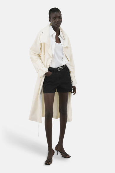 Zwarte bermuda short van MANGO, met een zilverkleurige gesp en gedragen met een witte blouse en trenchcoat.
