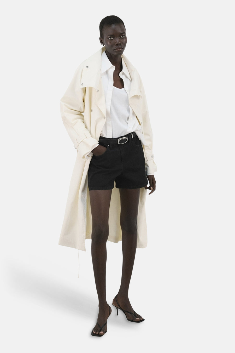 Bermuda short noire de MANGO, avec une boucle argentée et portée avec une blouse blanche et un trench-coat.