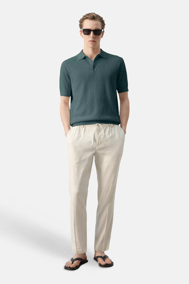 Polo côtelée verte de MANGO, à manches courtes, portée avec un pantalon clair, des lunettes de soleil foncées et des tongs noires.