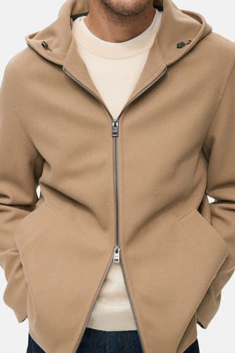 Manteau court beige de MANGO, avec capuche, fermeture éclair et poches, porté sur un gilet crème.