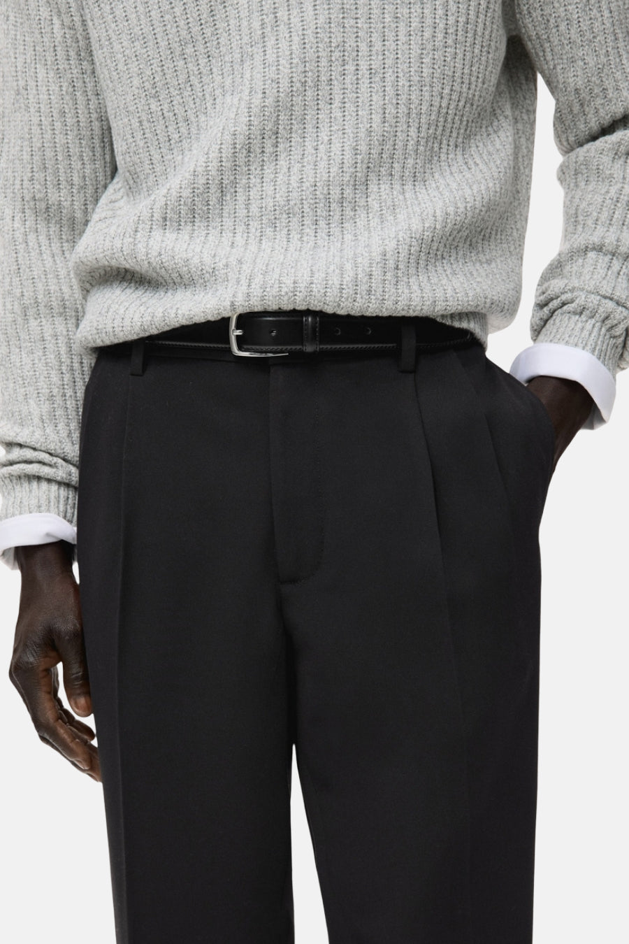 Pantalon noir de MANGO, avec plis et une fine ceine noire, associé à un pull gris.