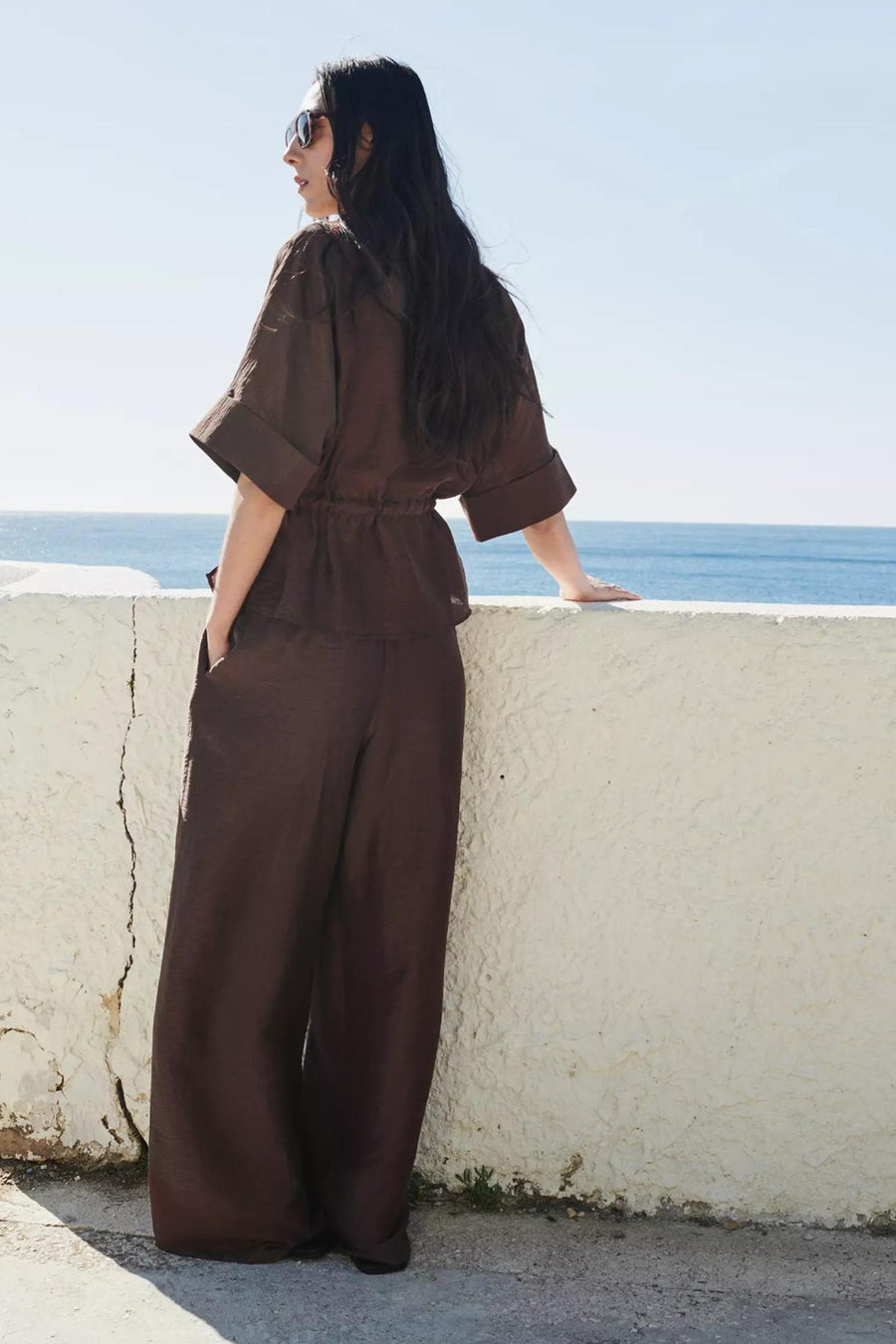 Bruine blouse met korte mouwen van MANGO, met trekkoord in de taille en opgerolde mouwen, staand aan zee.