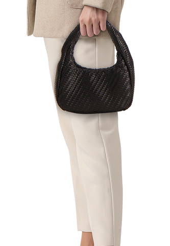 Schoudertas zwart van MANGO met gevlochten textuur, vastgehouden door een persoon in een beige jasje en crème broek.
