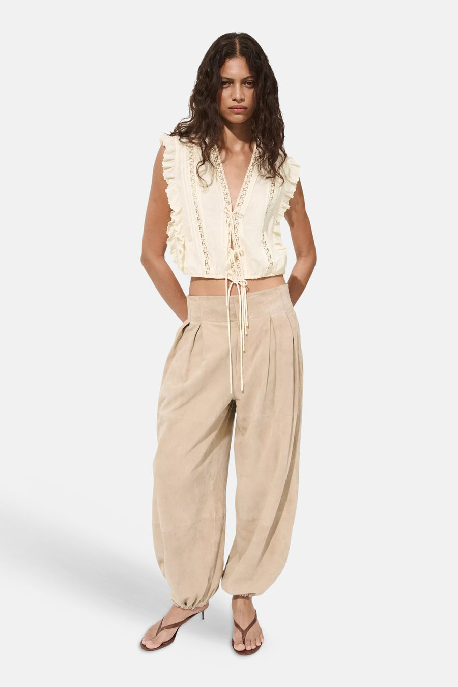 Beige mouwloze blouse met ruches en kant details van MANGO, voorzien van knoopsluiting en gekenmerkt door een diepe V-hals, gedragen met beige broek en sandalen.