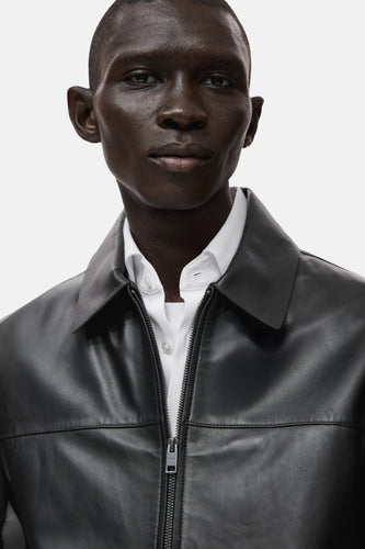 Zwarte korte jas van MANGO met een kraag en een ritssluiting, gedragen over een wit overhemd.