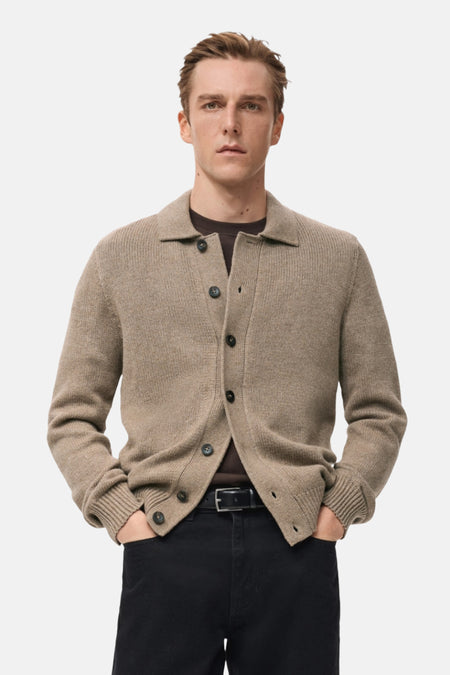 Man met bruin cardigan, zwarte broek, handen in de zakken.
