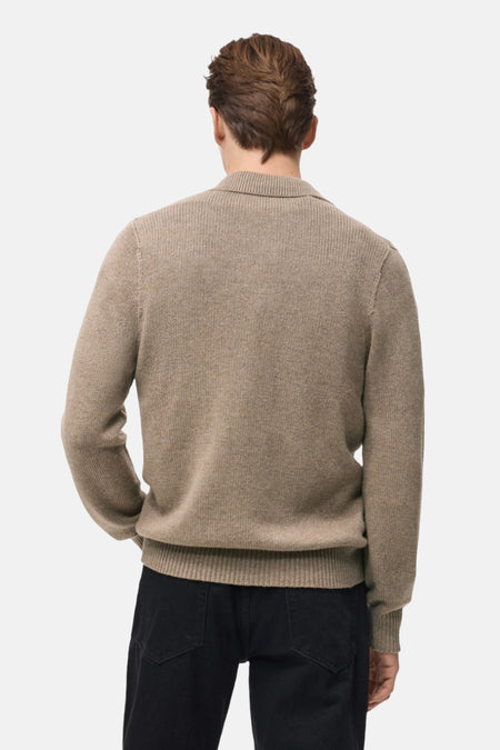 Achterkant van een man met een bruine wollen trui en zwarte jeans, staand tegen een witte achtergrond.