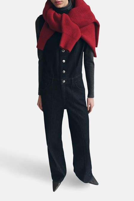 Donkerblauwe denim jumpsuit van MANGO, met zilverkleurige knopen en een rode gebreide trui nonchalant over de schouders.
