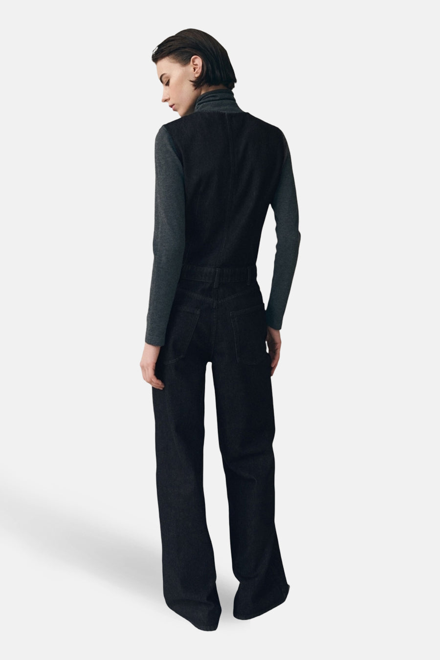 Donkerblauwe denim jumpsuit van MANGO, gezien van achteren, met lange, wijd uitlopende pijpen en een mouwloze top over een grijze coltrui.
