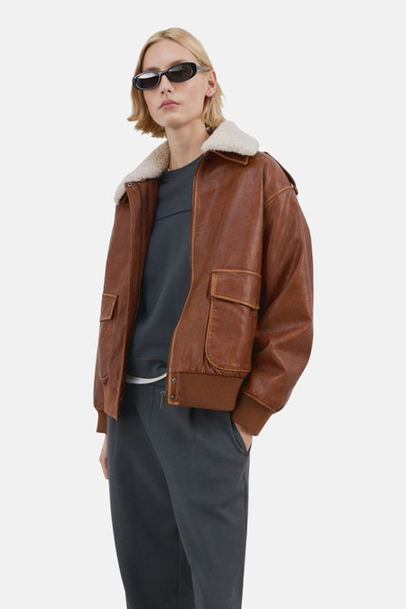 Pull-over bleu avec col rond de MANGO, porté sous une veste de pilote brune et un pantalon gris.