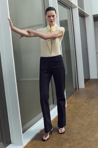 Gele mouwloze blouse van MANGO, met zwarte afwerking en open kraag, gecombineerd met een donkere broek en hakken.
