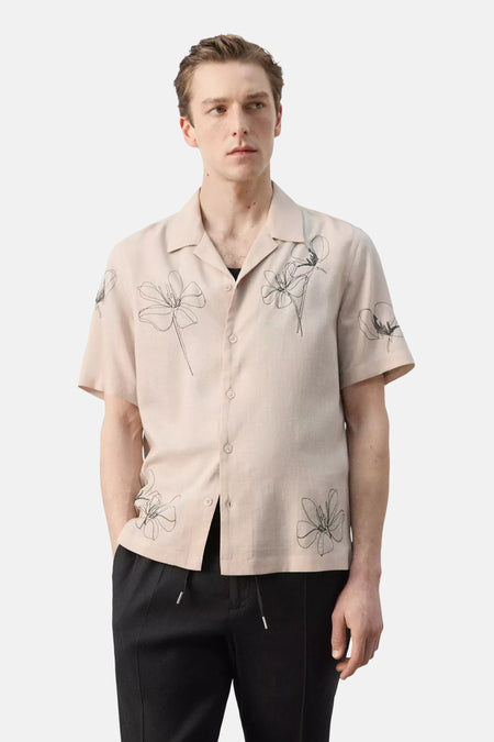 Chemise beige à manches courtes de MANGO, vue arrière, avec des fleurs brodées sur les manches, portée avec un pantalon noir.