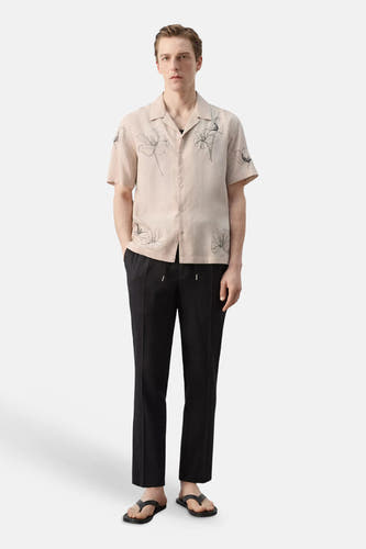 Beige overhemd met korte mouwen van MANGO, met een zwart bloemenpatroon, gedragen met een zwarte broek en slippers.