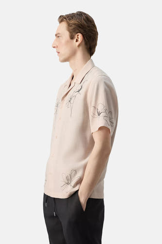 Zijaanzicht van een beigekleurig hemd met korte mouwen van MANGO, met zwarte bloemmotieven en Cubaanse kraag, gedragen door een man in een zwarte broek.