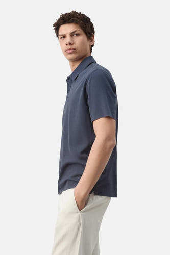 Blauwe polo van MANGO met korte mouwen en kraag, gedragen door een man met hand in de zak, zijzicht.