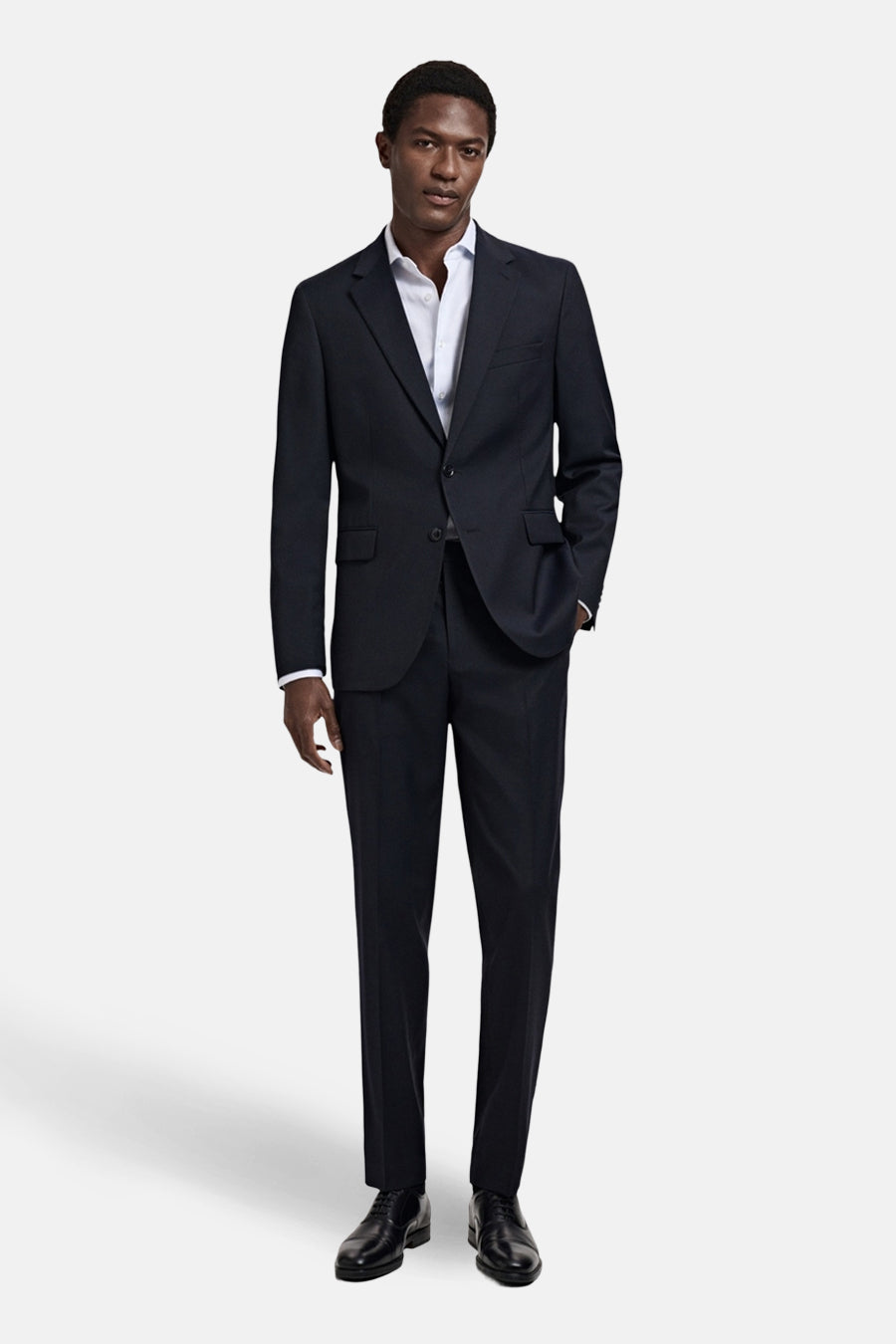 Blazer bleu de MANGO, avec une chemise blanche ouverte et des chaussures en cuir noires.