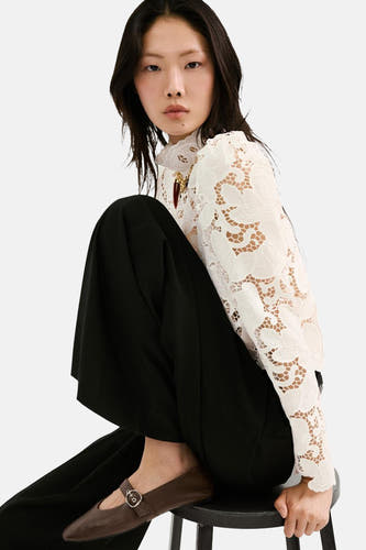 Chemisier blanc à manches longues de MANGO, avec un motif floral en dentelle, porté avec un pantalon noir et des ballerines marron.