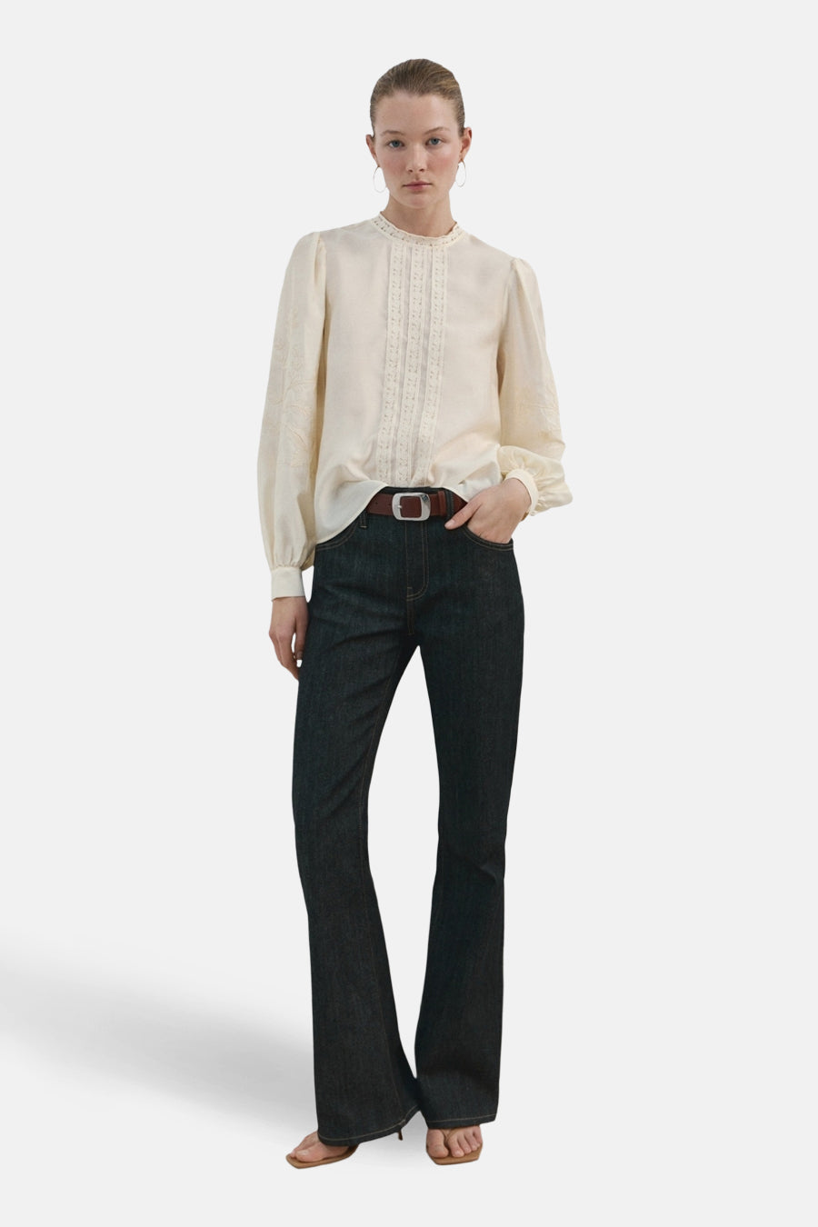 Blouse écru (manches longues) de MANGO, avec détails brodés le long du col et des manches, combinée avec un jean et une ceinture marron.