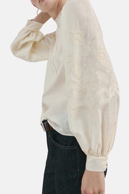 Ecru blouse (lange mouwen) van MANGO, met geborduurde bloemmotieven op de mouwen, gecombineerd met een donkere spijkerbroek en bruine riem.

