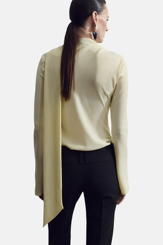 Gele blouse met lange mouwen van MANGO, met een lange sjaalkraag en zwarte pantalon.
