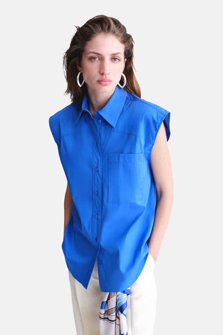 Blauw mouwloos hemd van MANGO met kraag en borstzak, gecombineerd met een witte broek en geprinte sjaal.