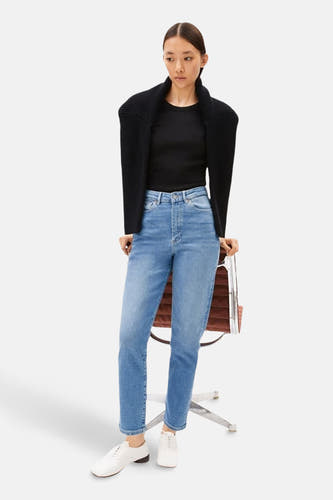 MAIRAA mid blue denim mom jeans van ARMED ANGELS, gecombineerd met een zwarte top en vest, en witte schoenen.
