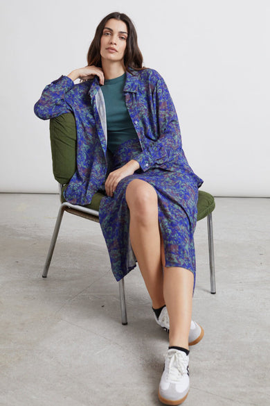 Chemise bleue à manches longues de 24 couleurs, avec un motif coloré, portée sur une chemisier verte et une jupe coordonnée.