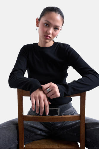 T-shirt noir à manches longues de TOPSHOP, porté par un modèle avec des boucles d'oreilles et une bre, assis sur une chaise en bois.