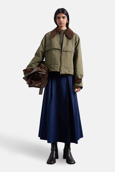 Manteau court vert de TOPSHOP, avec des détails marrons sur la en et les manches, porté avec une longue jupe bleue et des bottes marrons foncées.
