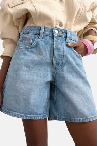 Lichtblauwe denim shorts van TOPSHOP, hoge taille, gewassen look, met hand in zak, beige overhemd en gekleurde armbanden, close-up voorzijde.