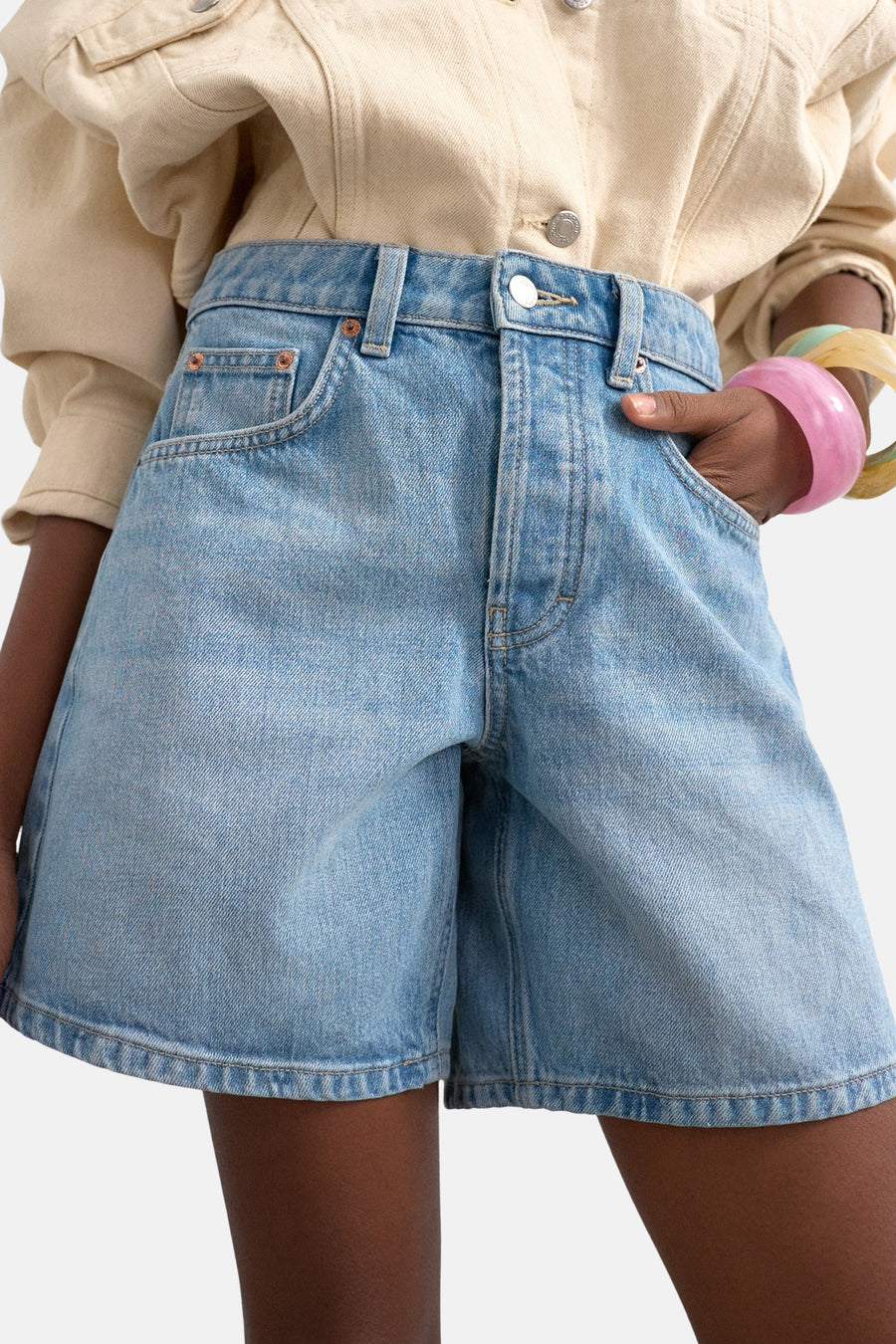 Lichtblauwe denim shorts van TOPSHOP, hoge taille, gewassen look, met hand in zak, beige overhemd en gekleurde armbanden, close-up voorzijde.