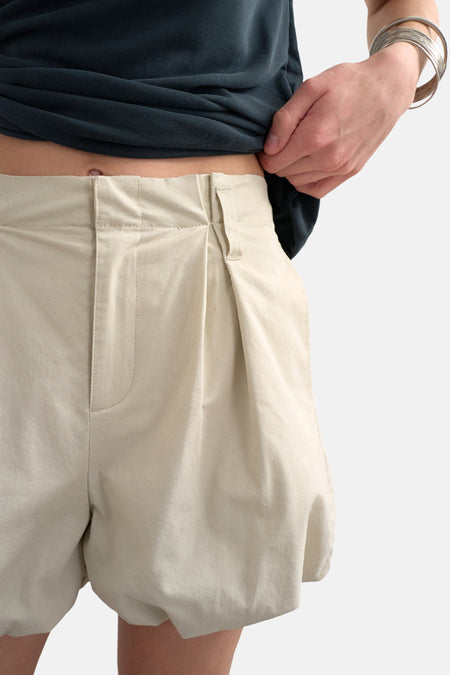 Ecru shorts van TOPSHOP met plooien, gedragen door een persoon die een donkerblauwe top optrekt en zilveren armbanden draagt.