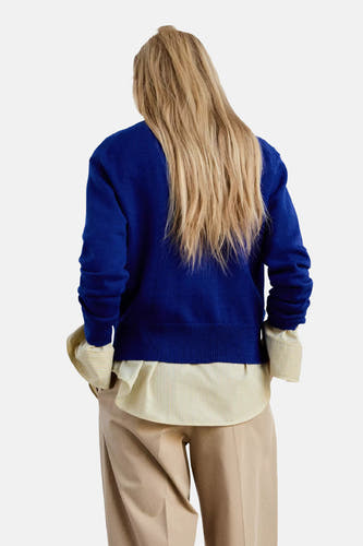 Blauwe V-hals trui van MANGO, achterzijde, met ribboorden en model in beige broek en gestreept overhemd