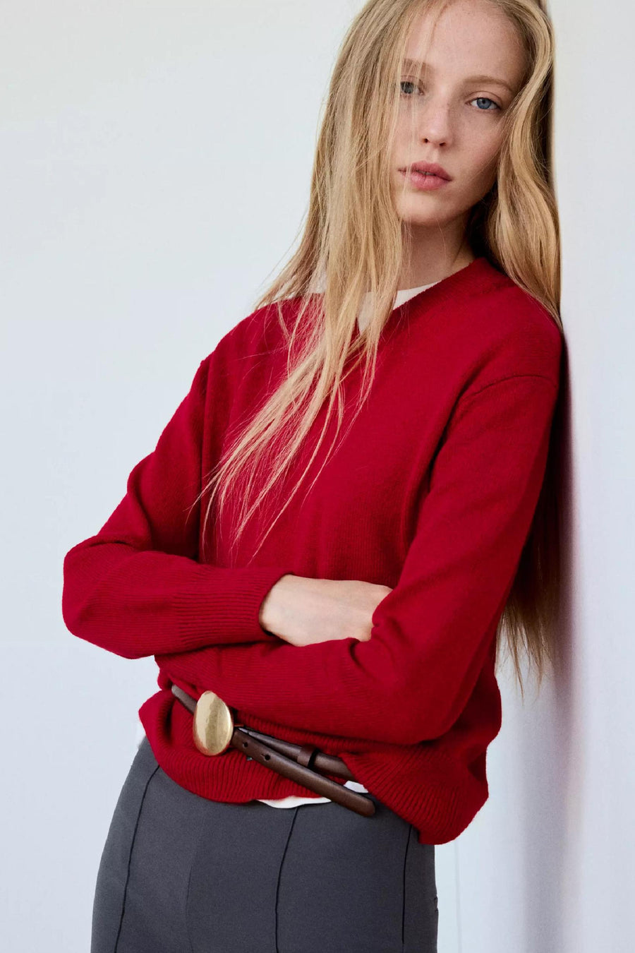 Rode V-hals trui van MANGO, gedragen met gekruiste armen, gecombineerd met een bruine riem en grijze broek, lifestyle shot.