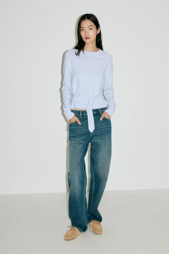 Lichtblauwe ronde hals trui van MANGO met riemdetail, gedragen met blauwe jeans en bruine schoenen, front view.