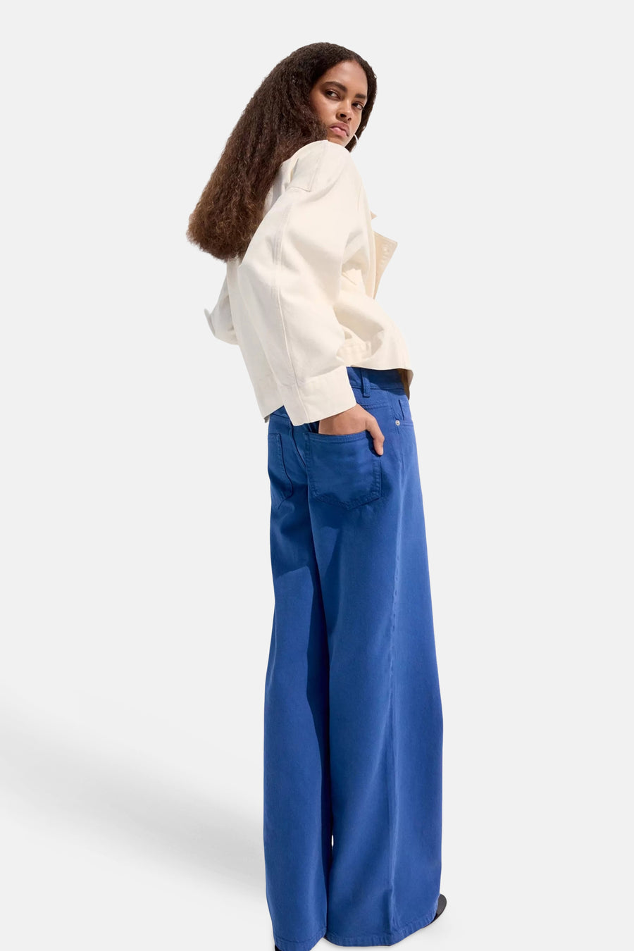 Blauwe damesbroek met wijde pijpen van MANGO, zijwaarts gefotografeerd, met witte blouse en hand in achterzak