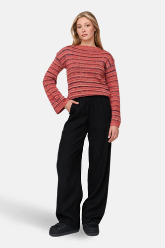 Jonge vrouw met een gestreepte oranje trui en zwarte wijde broek, staand tegen een witte achtergrond.
