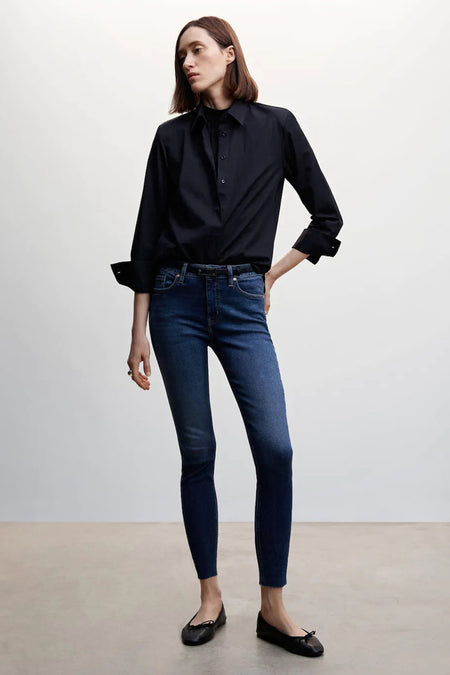 Jean skinny bleu de MANGO avec ourlets bruts, stylisé avec un chemisier noir et des ballerines.