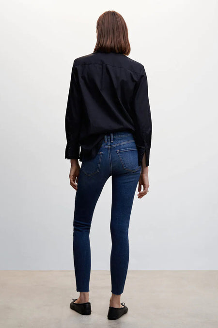 Middenblauwe skinny jeans van denim van MANGO met gerafelde zoom, gezien op een model van achteren, gecombineerd met een zwart overhemd en zwarte ballerina's.