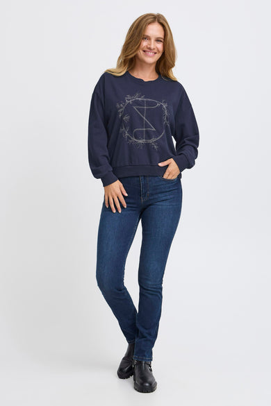 Blauwe sweater van PULZ, met decoratieve print op de voorkant, gecombineerd met jeans en zwarte laarzen.
