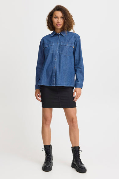 Jeanshemd in mid blue denim van PULZ, gedragen met een zwarte rok en zwarte boots.
