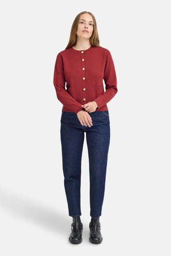 Rood gilet van PULZ, met lange mouwen en gouden knopen, gecombineerd met donkere jeans en zwarte laarzen.
