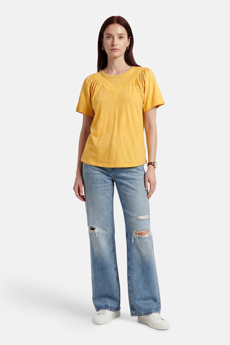 Chemisier jaune à manches courtes de PULZ, fini avec des détails en dentelle à l'encolure et porté avec un jean déchiré.