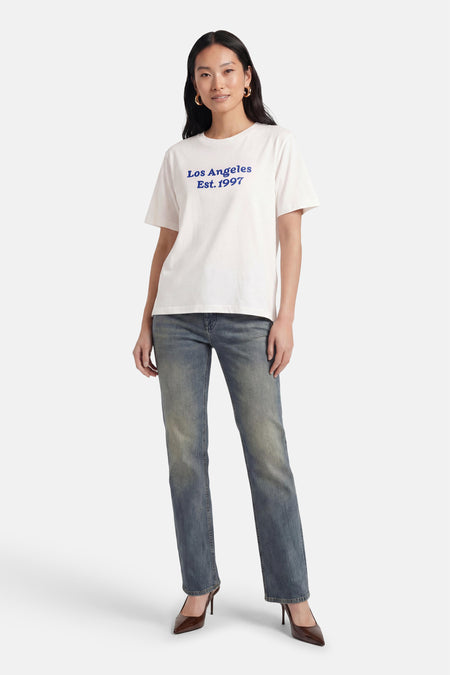 Wit T-shirt met korte mouwen van PULZ met blauwe tekst 'Los Angeles Est. 1997', gedragen door een model met spijkerbroek en bruine pumps.