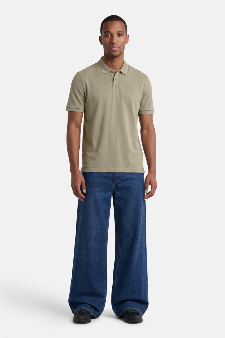 Polo verte HUGO verte de couleur verte avec logo et manches courtes, portée par un homme en jean large bleu foncé et baskets noires.