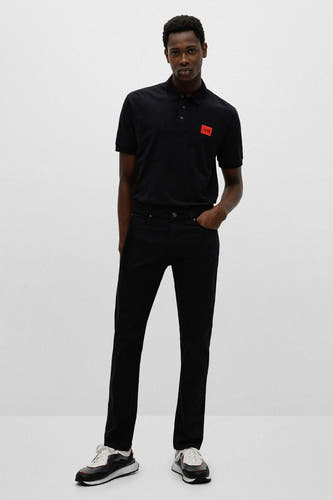 Zwarte polo van HUGO, met korte mouwen en een rood logo op de borst, gecombineerd met een zwarte jeans en sneakers.
