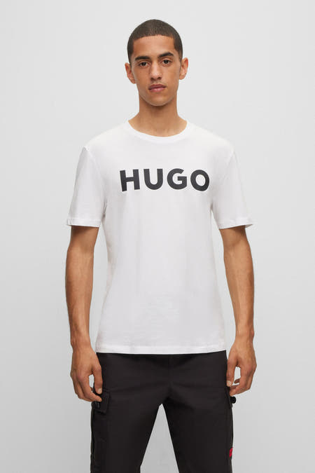 Wit T-shirt van HUGO, met korte mouwen en zwarte "HUGO" tekst op de borst, gedragen met zwarte broek.
