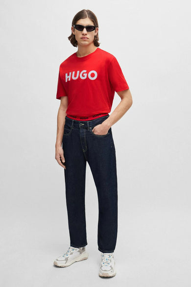 T-shirt rouge HUGO, avec une impression de logo blanc sur la poitrine, porté avec des jeans foncés et des baskets.