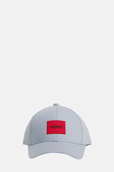 Blauwe pet van HUGO, met een rode merklogo-patch op de voorkant, en een lichtgekleurde knoop op de top.
