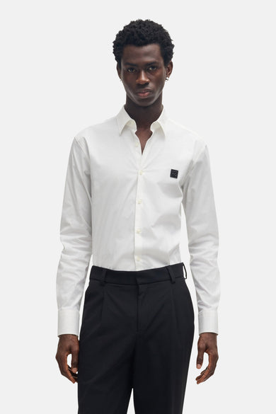 Chemise blanche à manches longues de HUGO, avec un logo foncé sur la poitrine et portée avec un pantalon noir.